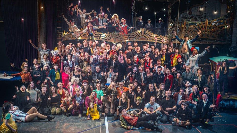 A massive group of performers in circus attire of all ages smiling with hands outstretched at the camera