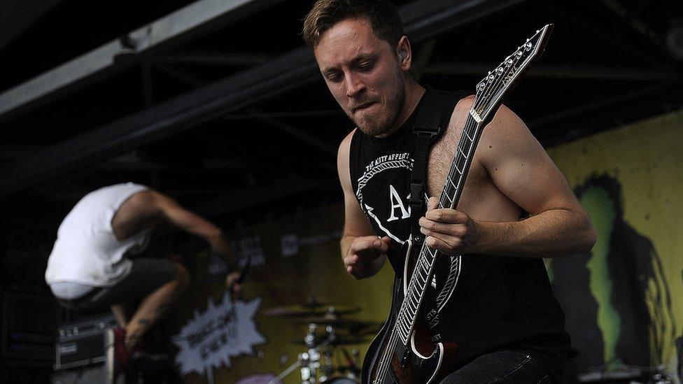 Tom Searle stands, playing the guitar on-stage with one knee supporting the instrument. He looks intense as he gears up to strum a chord - he appears to be gently biting his bottom lip as he concentrates. In the background a bandmate can be seen leaping into the air.