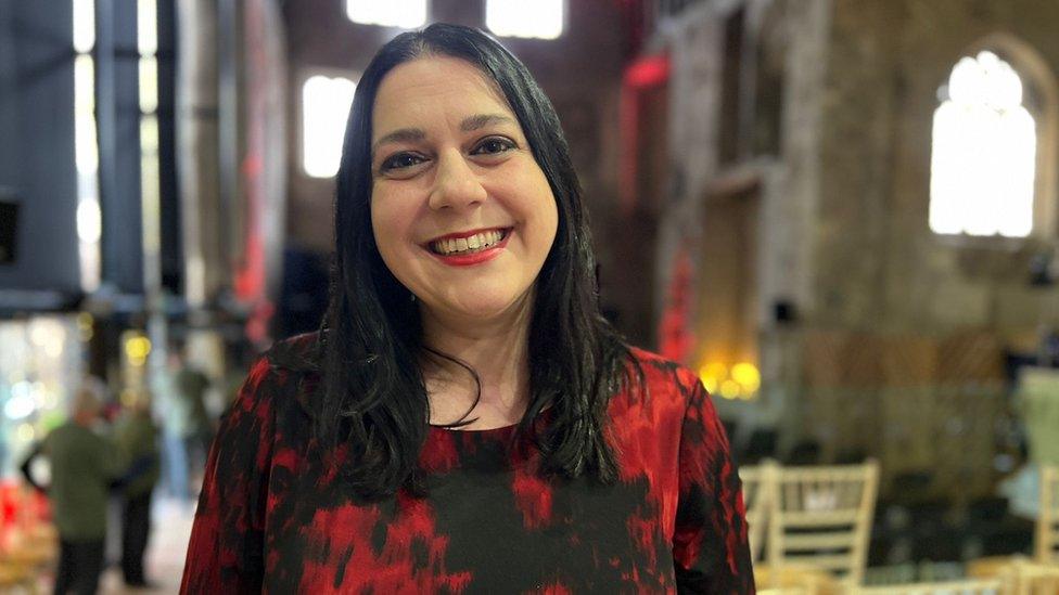 Dr Janina Ramirez in Gloucester Cathedral smiling while wearing a red and black top