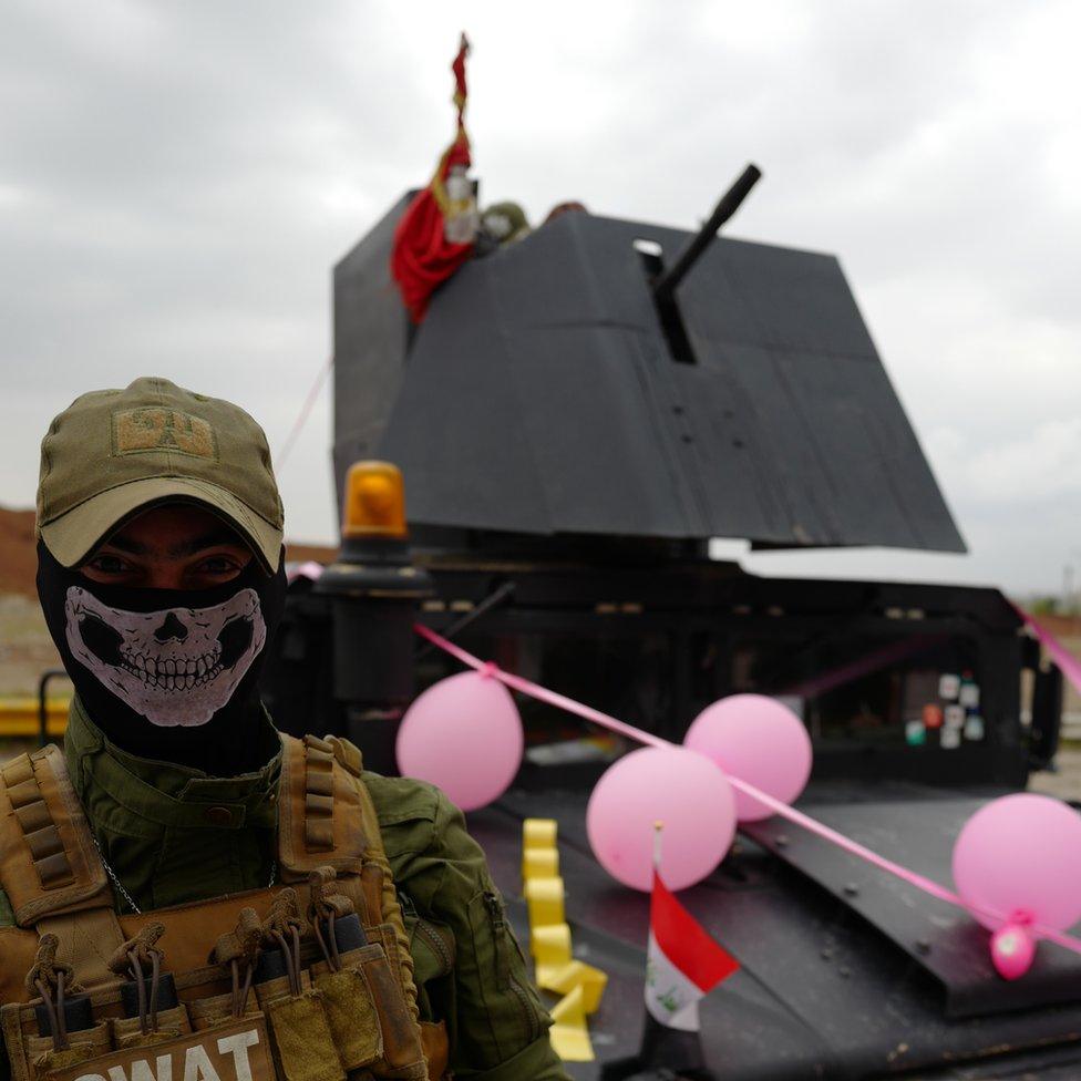 A SWAT officer with a skull mask over the lower half of his face stands in front of a military vehicle bedecked with pink balloons