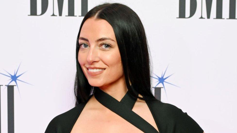 Jessica Agombar at the BMI Awards. Jessica is a young white woman with long dark hair worn loose over her shoulders and blue eyes. She smiles at the camera, her head slightly angled over her right shoulder. She wears a black dress with a strap over her chest. She is pictured in front of a white back drop with the BMI logo printed on it.