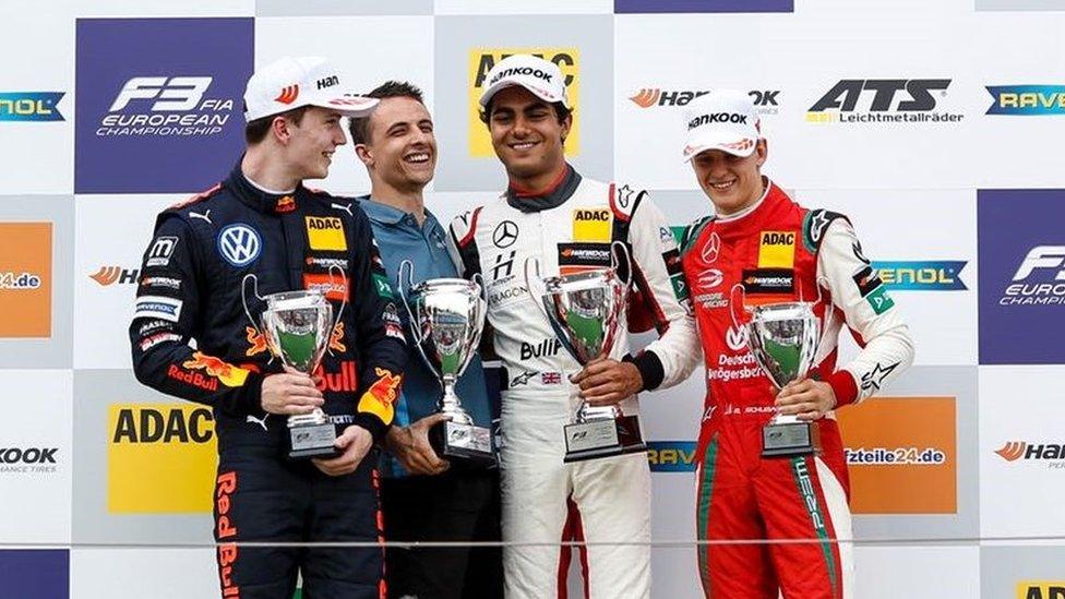 Enaam Ahmed is stood on a podium with three other racing drivers. He is standing second from the right wearing a white Mercedes racing driving overalls with a white cap. He is smiling and holding a silver trophy in his left hand. Stood next to him are three white racing drivers who are also all wearing racing overalls and holding silver trophies and smiling. Behind them is a sponsors board with various racing sponsors on it.