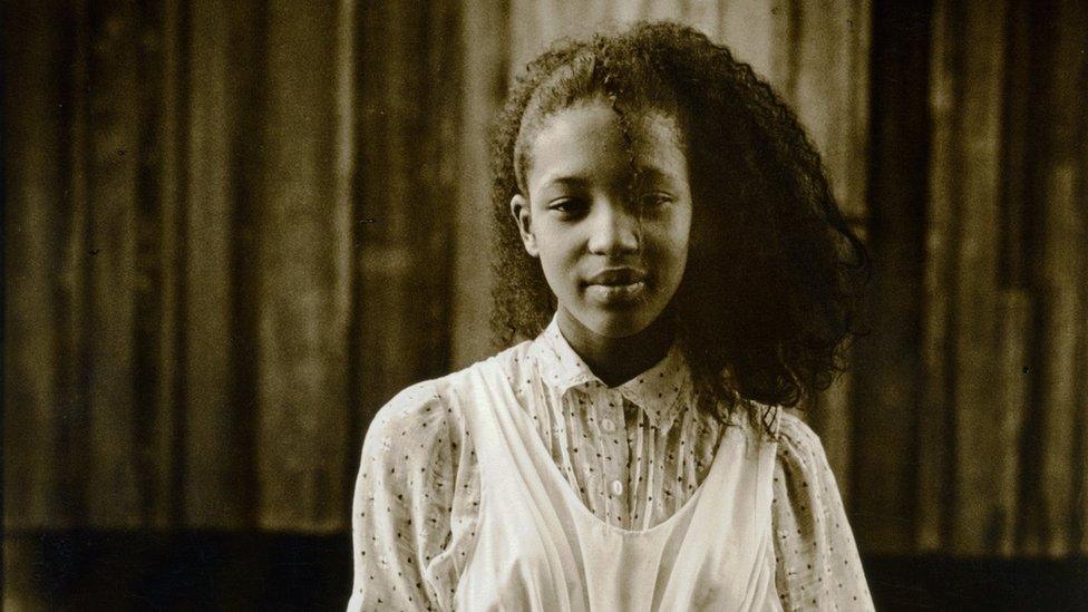 A black and white image of a young Naomi Campbell with long hair in a white dress and patterned shirt