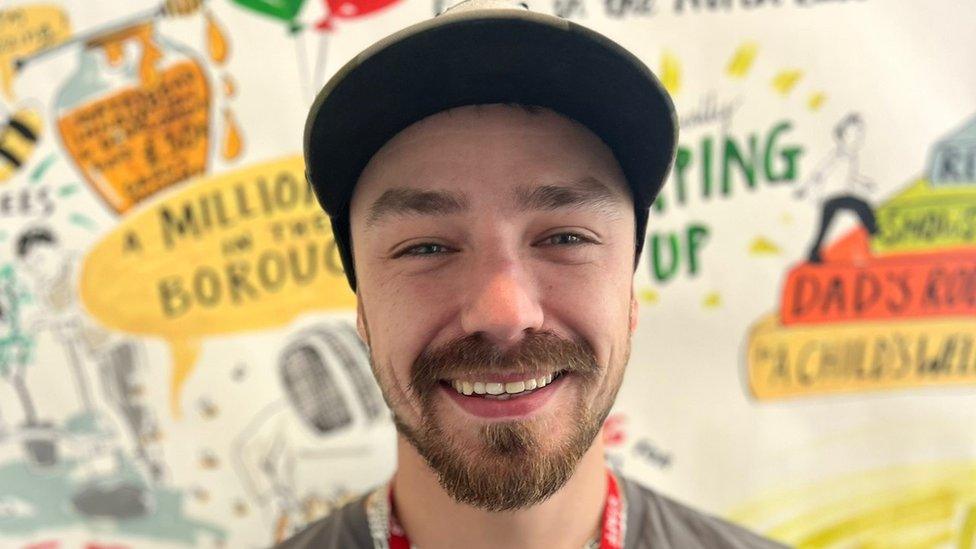 A photo of young dad Nathan who is smiling for the camera. He has a cap on with a black peak and has a dark goatee style beard. It's a close up photo of his head and behind him is a mural on the wall with positive phrases on about being a dad.