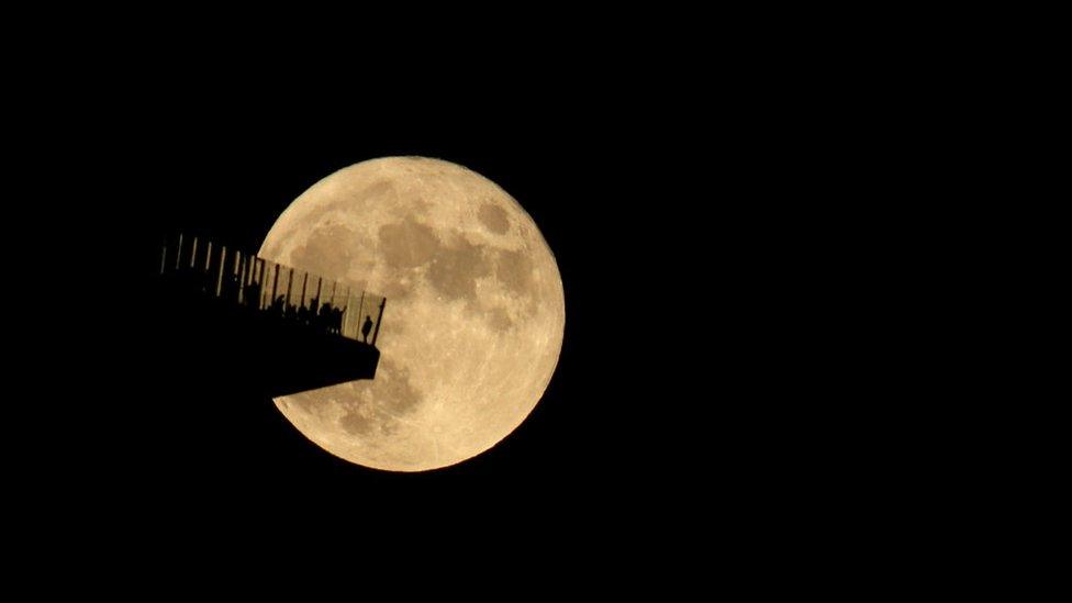 Silhouettes of people on a glass walled deck high up in front of a big moon