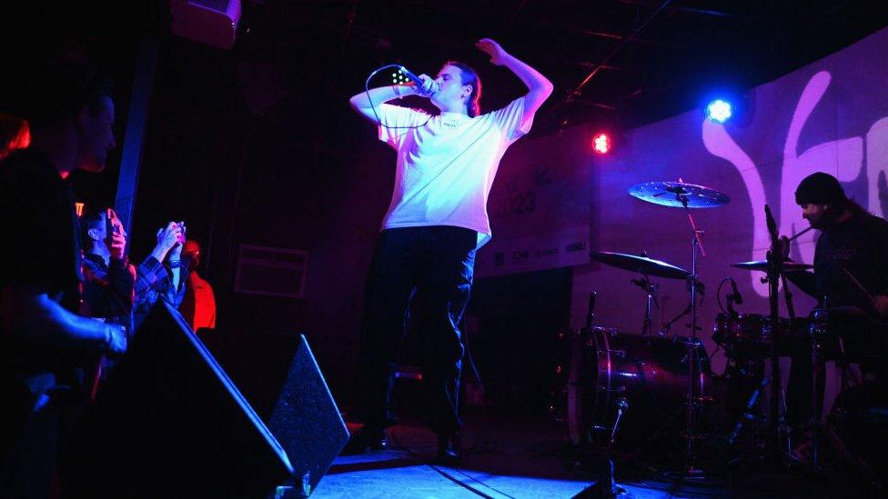 Venbee performing on-stage. She's wearing a white t-shirt and black trousers. She's bathed in a purple light, her eyes are closed and she has one arm, bent above her head while the other hand holds the microphone close to her mouth.