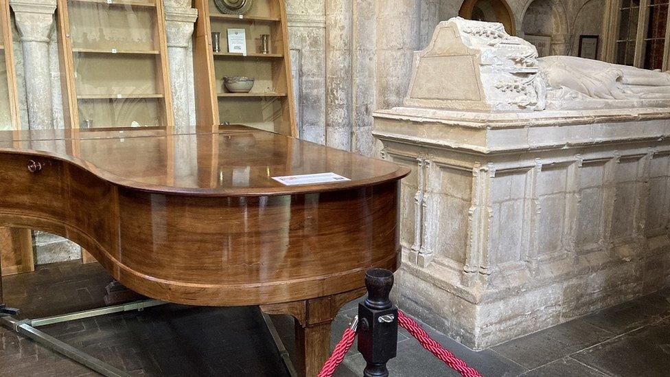 Corner of the abbey showing a large stone tomb on the right, a piano in the middle, display cases at the back and a piece of red rope at the front.