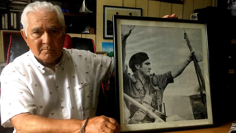 Edén Pastora holds a picture of himself with a rifle in his left hand