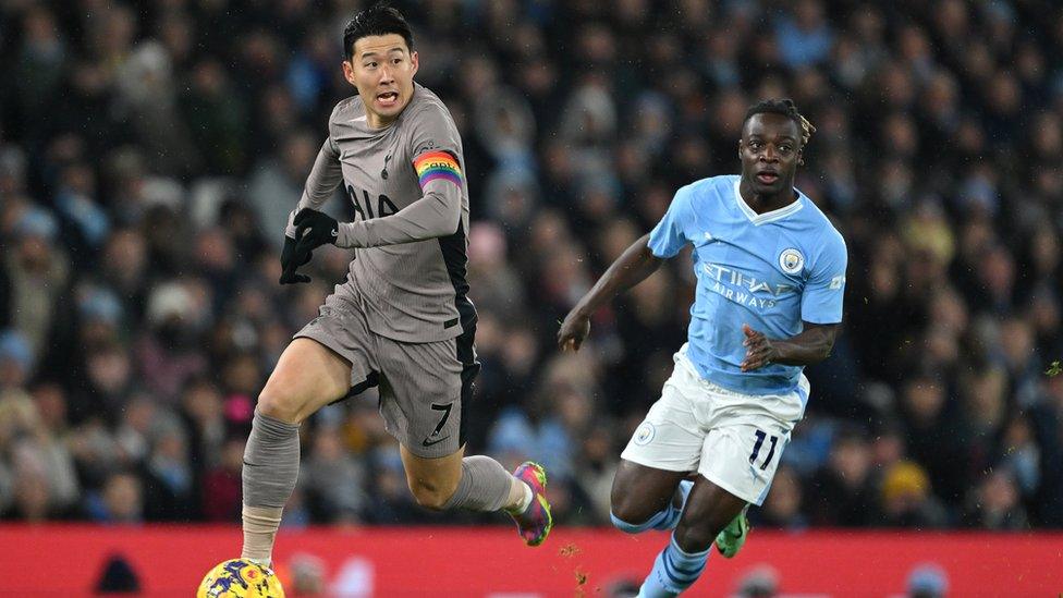 Son Heung-Min and Jeremy Doku playing football. Son is an Asian man and is dribling with the yellow football at his right foot. He is wearing a browny grey Tottenham Hotspur away kit which has a black stripe down the side. The socks and shorts are the same browny grey colour and he is wearing black gloves. He has short black hair and is wearing a captain's armband on his left arm which says "captain" and is the colours of the rainbow. Doku is a black man and he has dark hair which is tied back and is wearing a light blue Manchester City shirt, with white shorts and light blue socks. He is chasing Son so is behind him and to the right, and Son is looking up to his right.