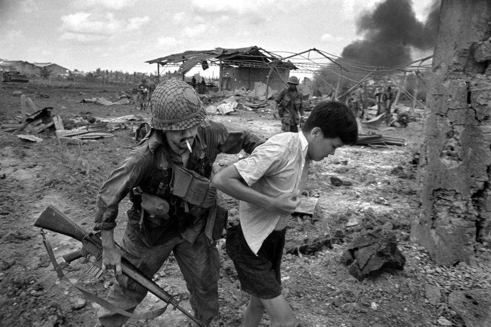 An American soldier forcefully pushes a Vietnamese person in a scene of devastation