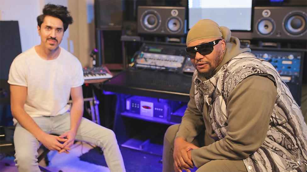 Mobeen Azhar and Malik Yusef. Mobeen is wearing a white tshirt and lightly coloured jeans, with his hands joined together as he looks at the camera. Malik is on the right, wearing sunglasses, a green bandana on his head, green hoodie and tracksuits with a grey checked covering over his top. The background has a table with speakers and music faders, and a neon blue light shining below the table.