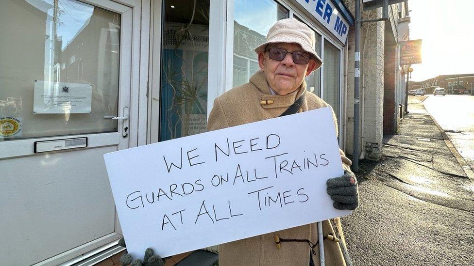 Mr Waddell holding a sign which says "we need guards on all trains at all times"