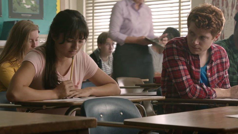 Two high school students sit in class as a boy looks across at the girl next to him.