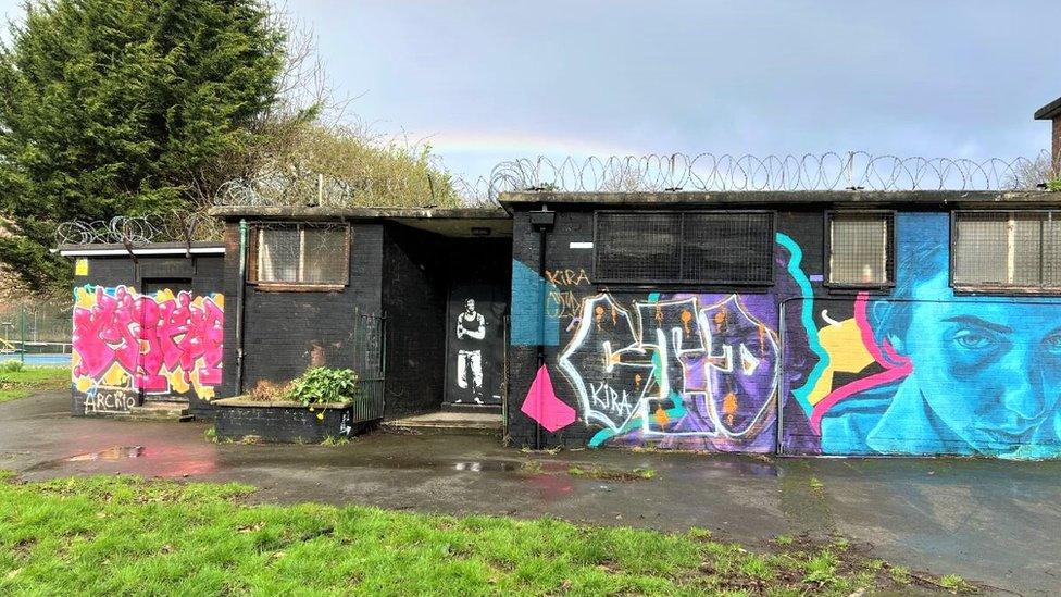 White spray painted mural of Bristol rapper Tricky on the black external wall of the bowling club. There are other colourful works of graffiti around it and a faint rainbow in the sky