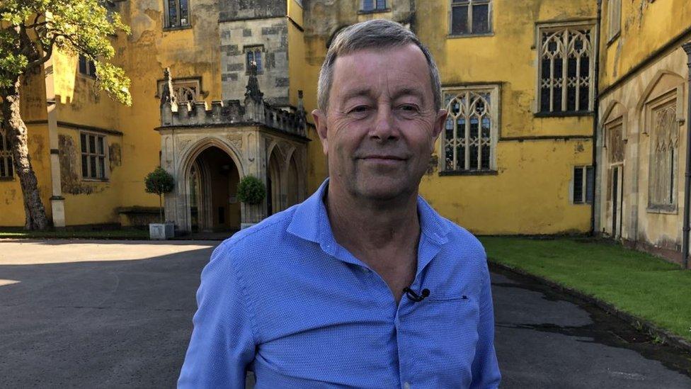 A middle aged man in a blue shirt in front of a yellow mansion