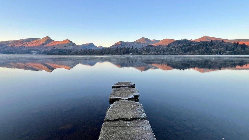 View across a lake with a few frosty rocks but a clear reflection of mountains in the background in the water