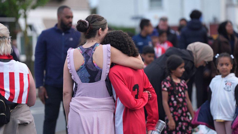 Members of the community, including River Hawks FC, looking at floral tributes in Hainault