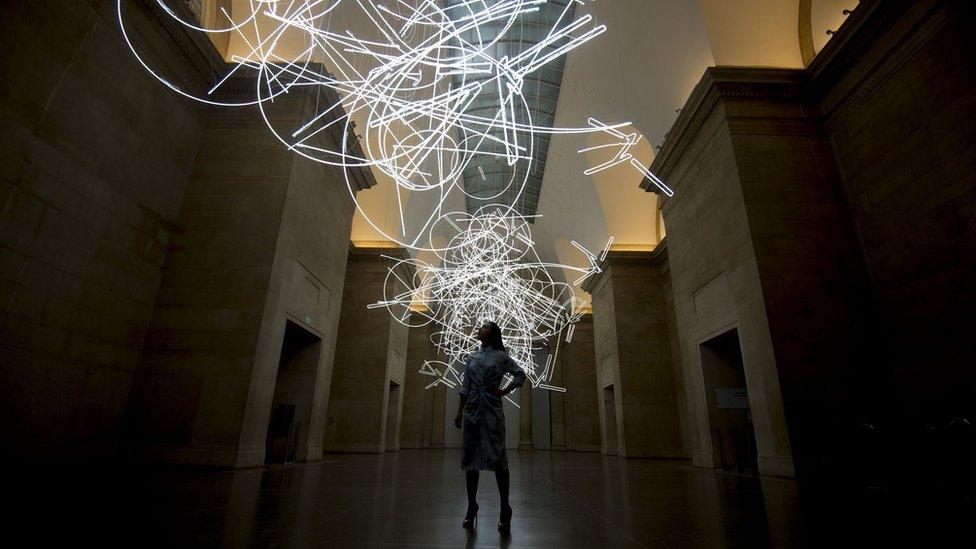 A gallery assistant stands under the neon sculpture