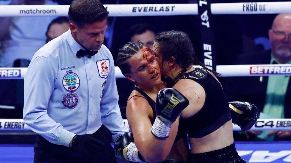 Two women with arms around each other in a boxing ring, with the referee looking on