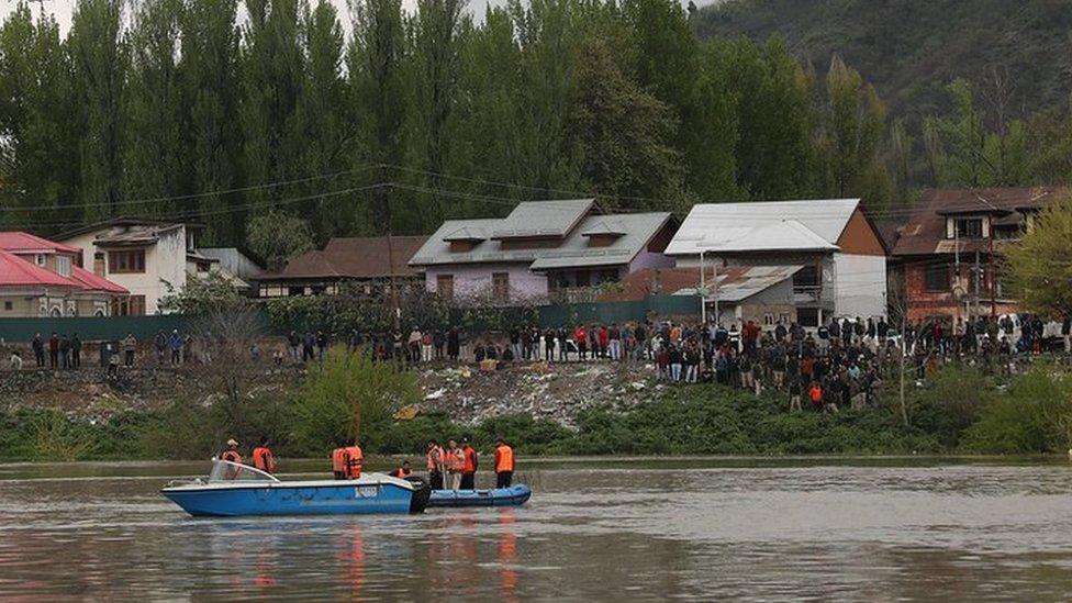 Onlookers stand on the river bank as rescuers in a boat search the water