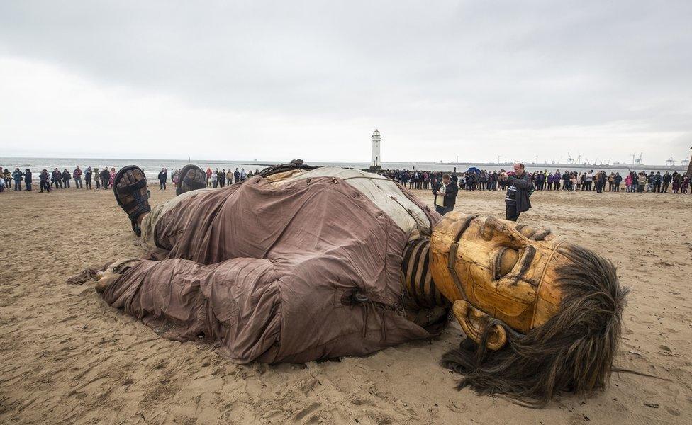 A giant puppet is seen on the beach surrounded by people looking on