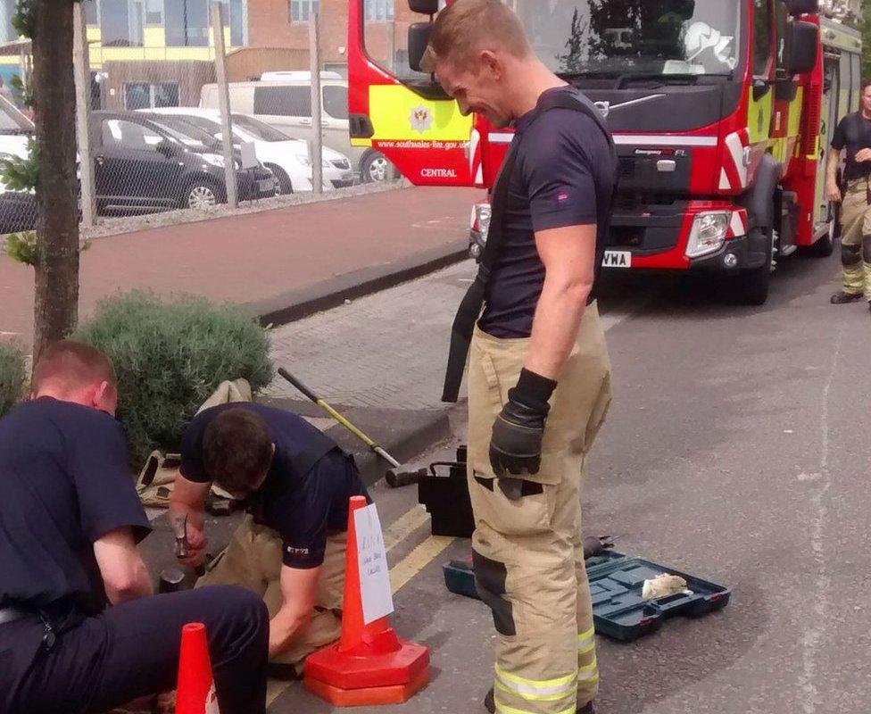 Three firefighters standing around a drain - one of them appears to be holding a crowbar and trying to open the drain. A fire truck is in the background