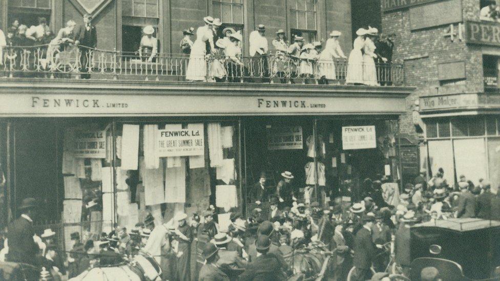 Fenwick family on store balcony with crowd passing in front