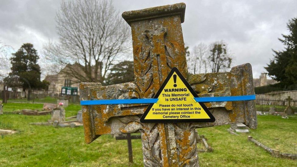 A cross headstone with a yellow warning taped onto it with blue tape. The warning reads: "Warning! This memorial is unsafe. Do not tamper with temporary support. Please contact Cemetery Office."