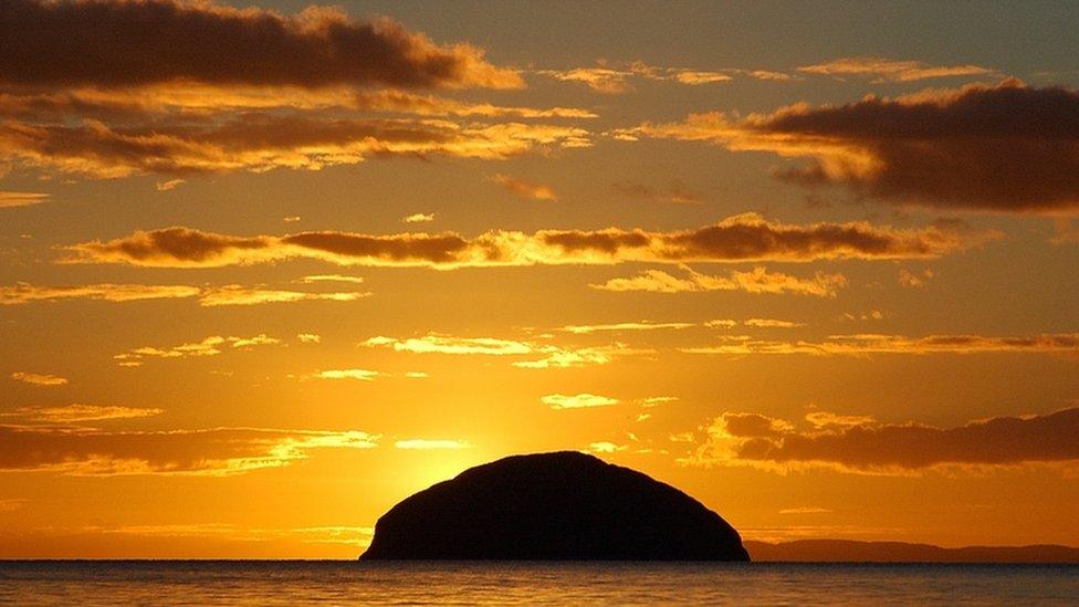 Bright orange and yellow sunset with some scattered clouds with the silhoutte of the Ailsa Craig