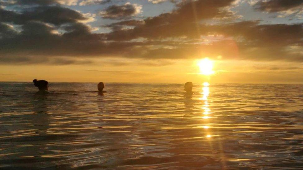 People swimming in the sea as the sun off Scarborough, as the sun sets in the background, its rays shimmering on the calm water