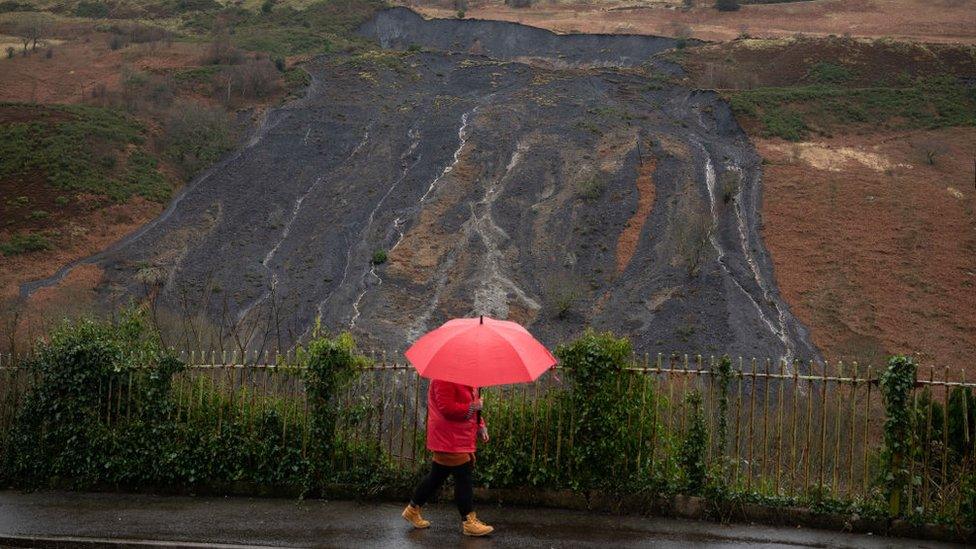 Woman with umbrella walks past coal tip landslide in Tylorstown, Rhondda Cynon Taf