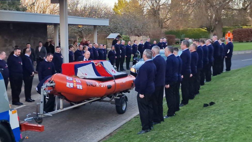 People in dark clothes lining the path while a lifeboat with a coffin on top is driven past