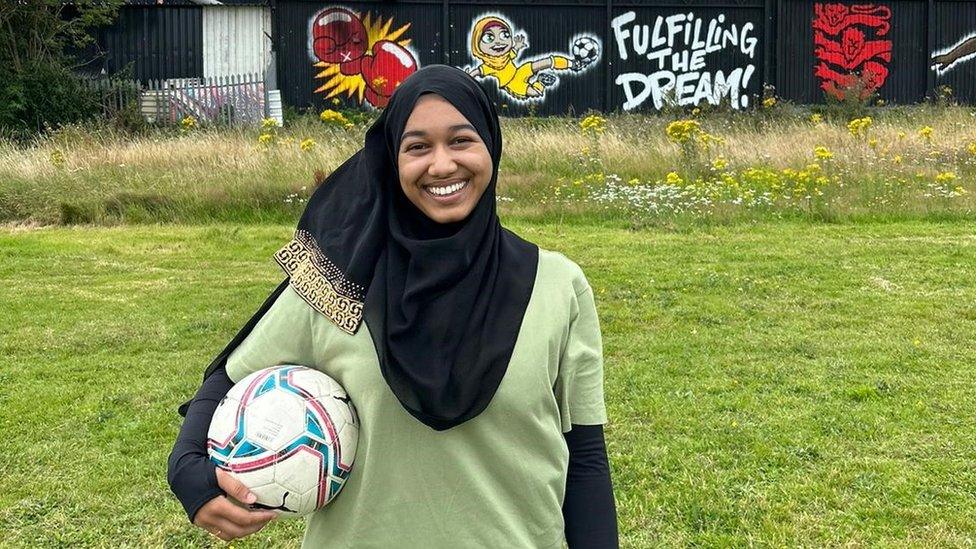 Yasmin Rahman standing in a grassy field holding a white football under her right arm. She is wearing a black hijab (headscarf) on her head and is smiling. She is wearing an olive green short-sleeved T-shirt and has a black long-sleeved shirt on underneath that. Behind her is a building with a mural on it that says "fulfilling the dream".