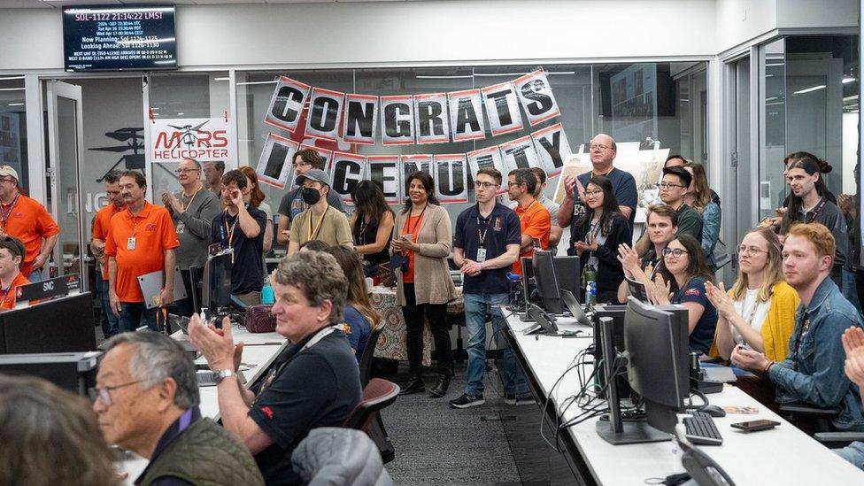 People sitting in an office all looking in one direction with a 'Congrats Ingenuity' sign in the background