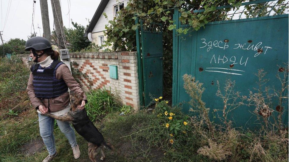 A journalist stands outside a garden wall and a gate with the inscription, "People live here"