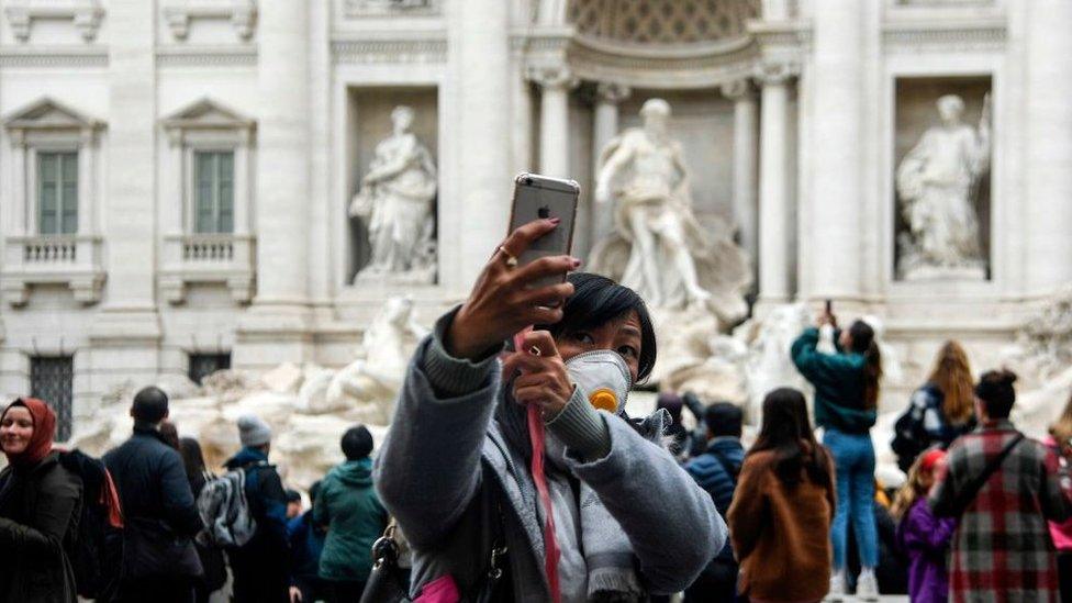 A Chinese tourist by the Trevi fountain in Rome, taking a selfie while wearing a face mask