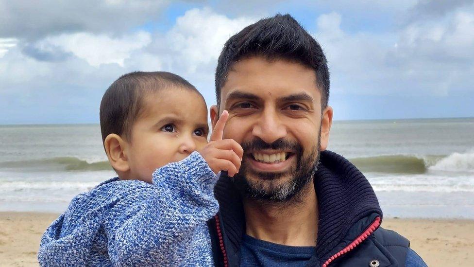 Avnish wearing a navy blue jumper standing in front of a beach and ocean, holding his baby son who is wearing a blue jumper and pointing up to the sky. They are on a beach with the ocean water behind them and a cloudy sky.