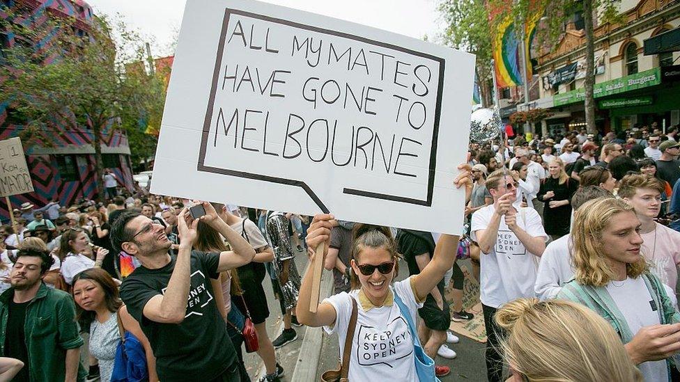 A woman at a protest holds a sign saying "all my mates have gone to Melbourne"