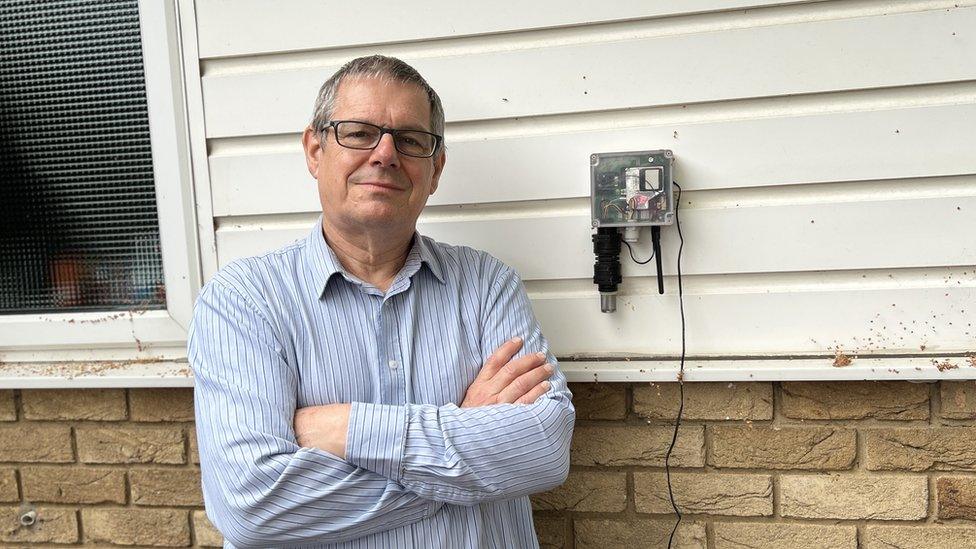 Man with short hair and glasses stands with arms folded next to small gadget.