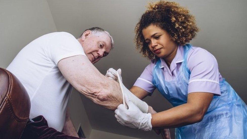 A carer puts a bandage on an old man's arm while he sits on a sofa