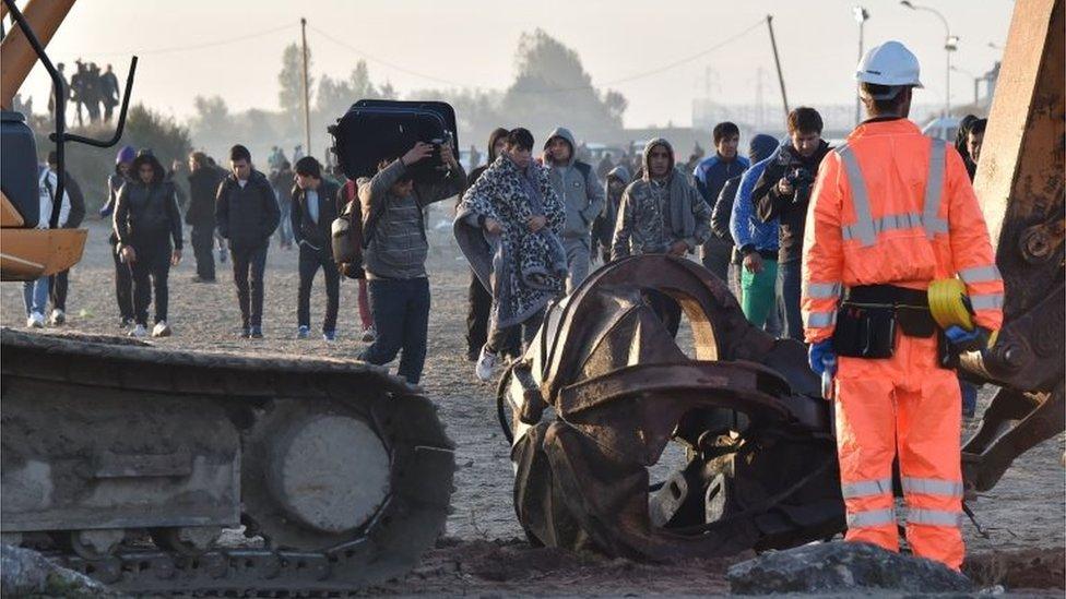 Migrants walk by excavators and Members of the demolition crew in "Jungle" migrant camp in Calais, northern France, on 27 October 2016