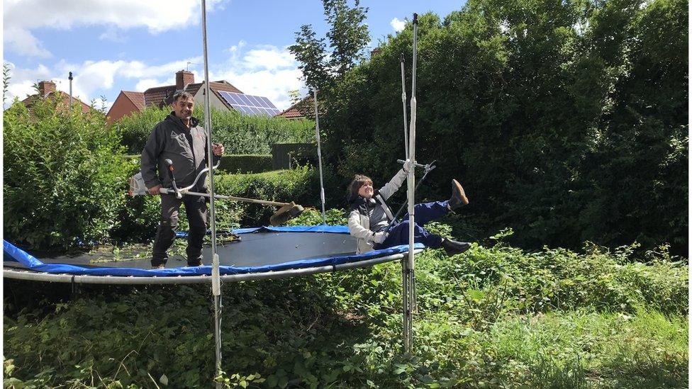 John with Bill's son on top of a trampoline, holding gardening tools. The pair are surrounded by overgrown hedges.