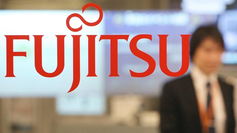 Fujitsu logo is seen pictured on glass with an out-of-focus employee in the background, taken at the company's offices in Tokyo in 2007