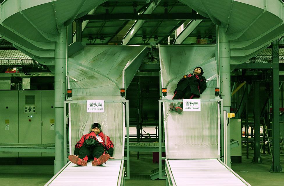 A photo by Cao Fei showing two people sliding down an industrial shoot in a warehouse