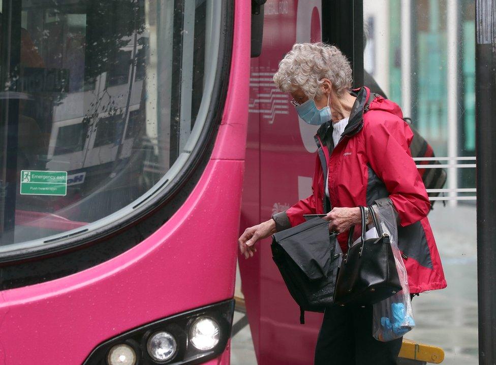 An elderly woman wearing a mask gets on to a bus