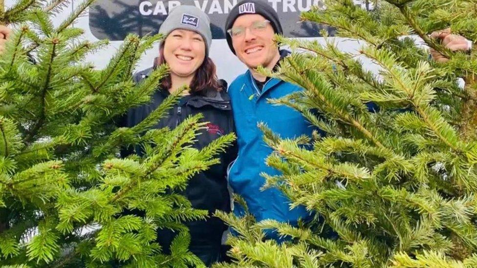 Two volunteers with a tree each in front of a van, smiling at the camera