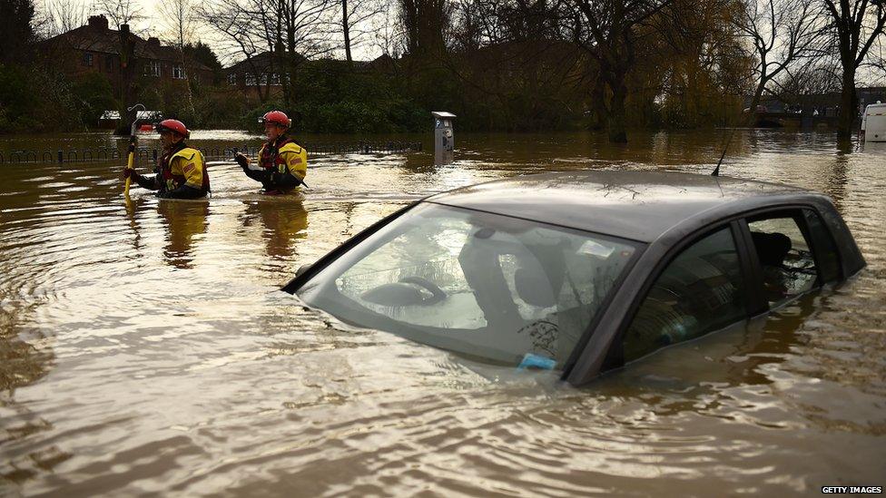 Car surrounded and nearly covered by flood water. Two rescuers behind, wading through the water.