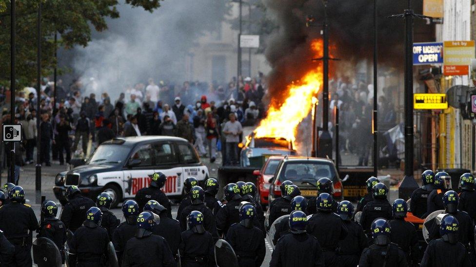 Police officers in riot gear block a road near a burning car on a street in Hackney, east London August 8, 2011