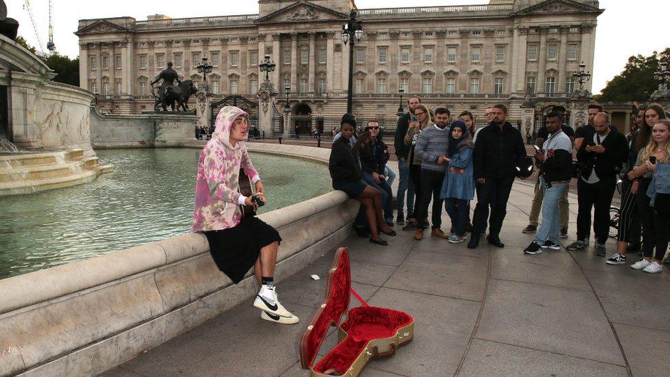 Justin Bieber busks while sitting on the edge of a fountain outside Buckingham Palace, while fans watch on