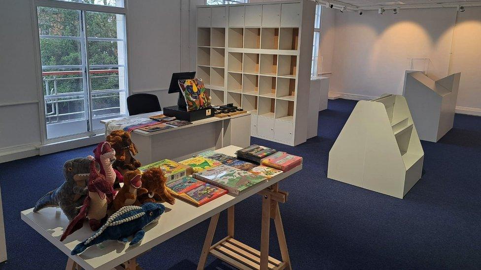 Image inside the new museum space. Empty shelves and a table full of books and stuffed toys can be seen.